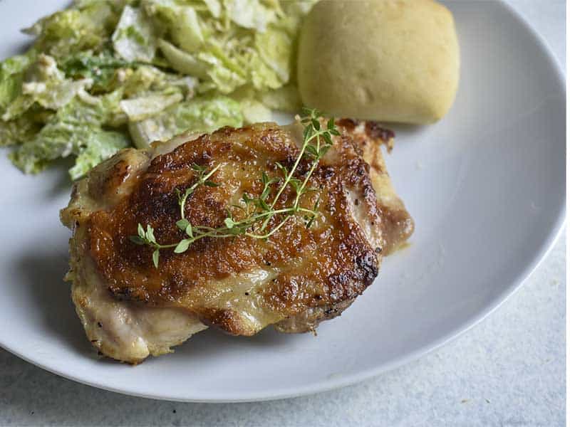 sous vide chicken thigh with crispy skin on a white plate with a bread roll and ceasar salad