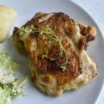 sous vide chicken thigh with crispy skin topped with fresh thyme next to a roll and ceasar salad.