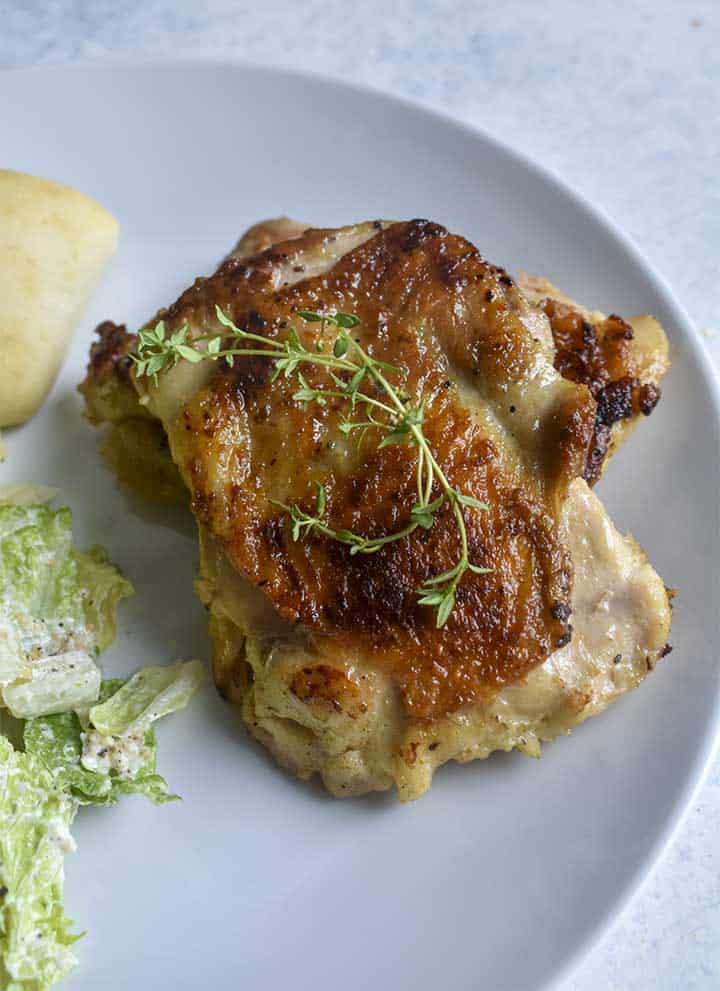 Sous Vide Chicken Thighs with golden brown skin on a white plate sitting on a white table. Topped with fresh thyme alongside cesar salad and a breadroll