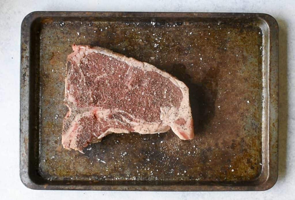 bone in ribeye steak seasoned with salt and pepper sitting on a rusted pan on a white table