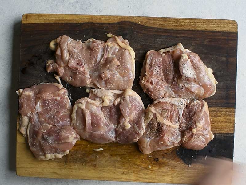 chicken thighs seasoned with salt and pepper upside down on a wooden cutting board on a white table