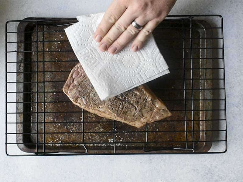 gray cooked steak being patted with a paper towel on a wire rack sitting over a rusted baking sheet on a white table