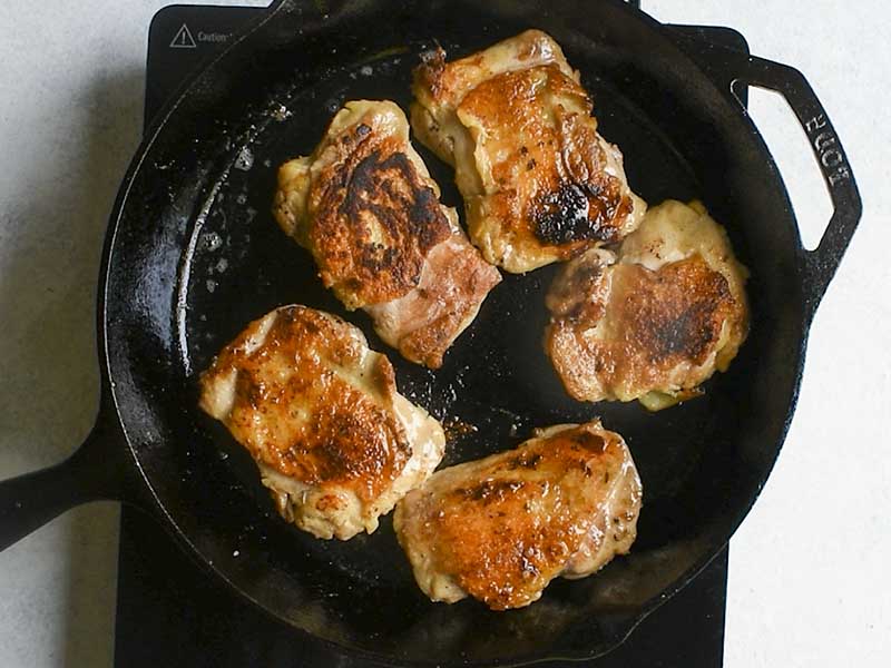 chicken thighs with a golden brown skin cooking in a black cast iron pan on a black electric griddle sitting on a white table