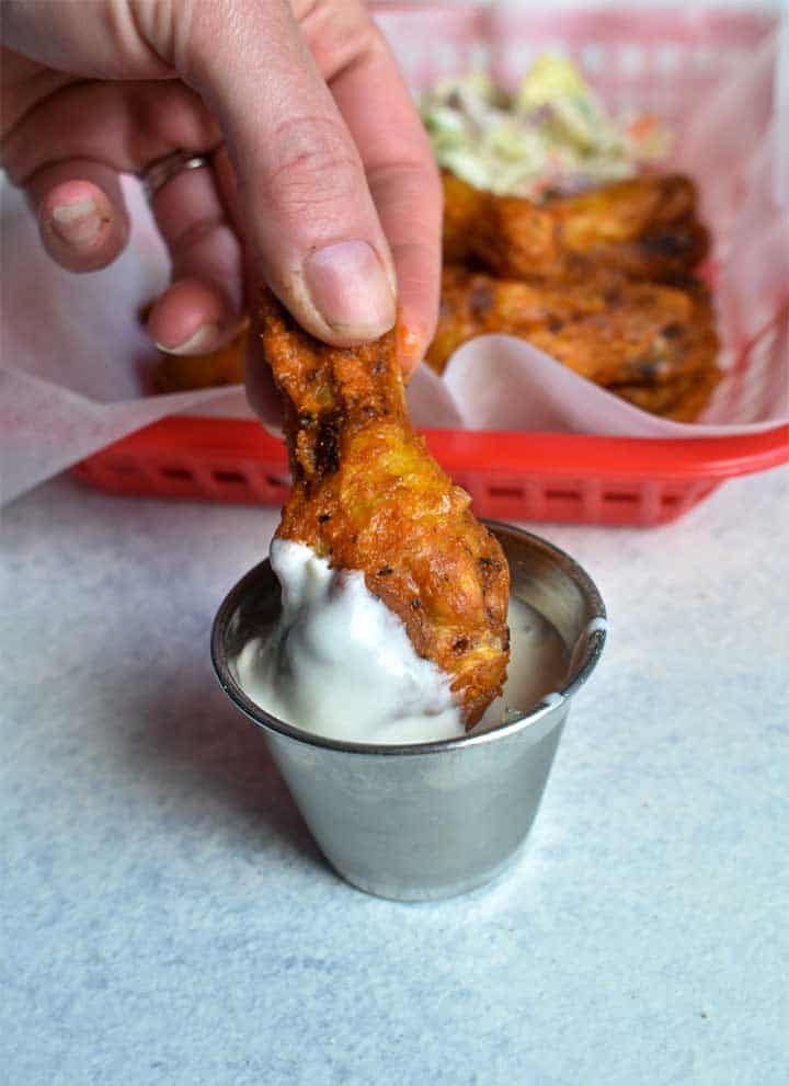 woman dipping chicken wings into blue cheese dressing in a metal dipping container on a white table. Chicken wings and salad sitting in a red basket with food service pepper in background.