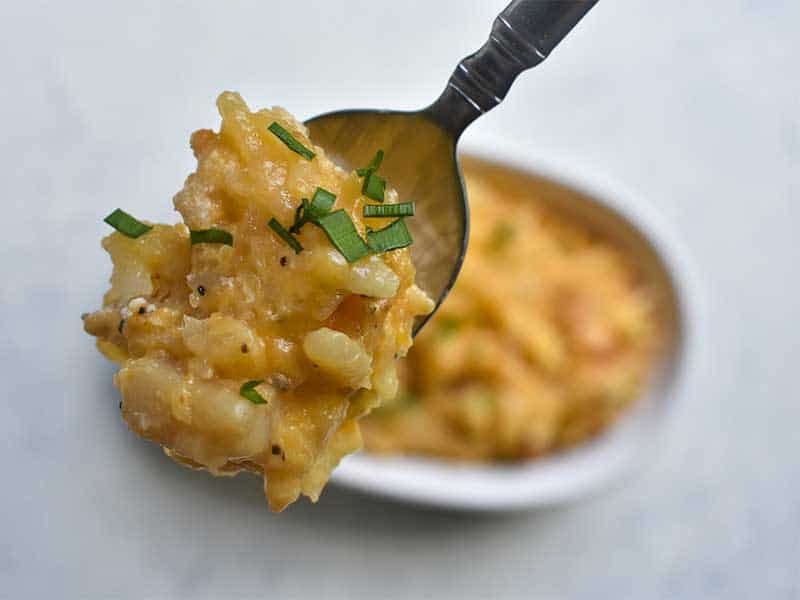 cheesy potatoes on a silver metal spoon above cheesy potatoes sitting in a white oval dish on a white table 