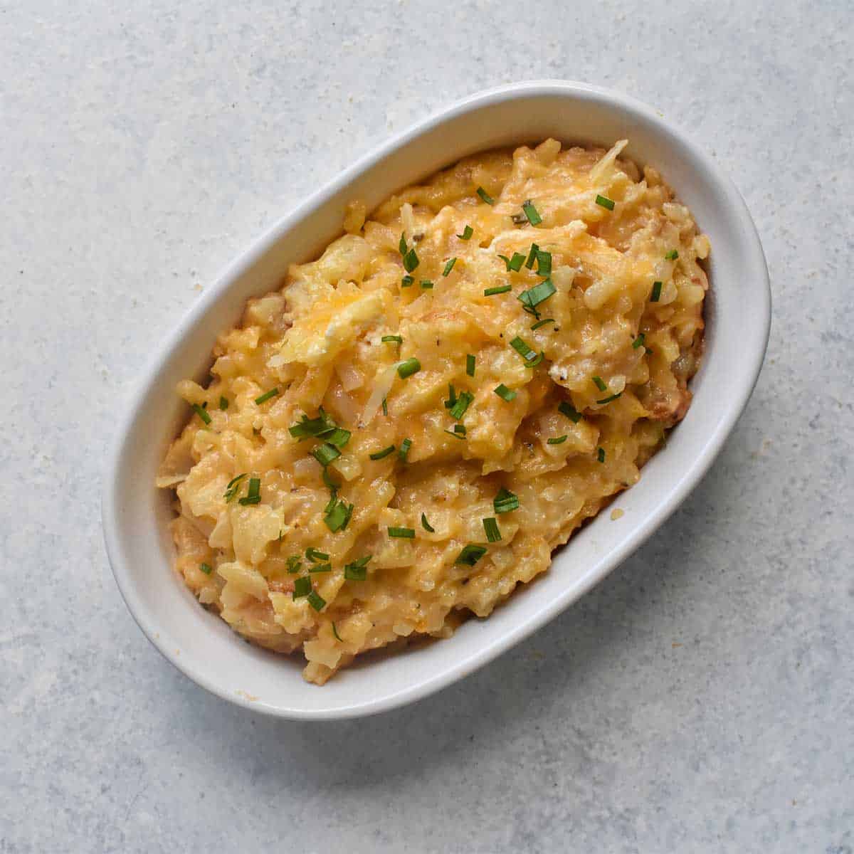 cheesy potatoes garnished with chopped chives in an oval shaped white serving dish on a white table