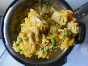 a large serving spoon with chicken and yellow rice with red peppers and peas above an instant pot which is blurry in the background.