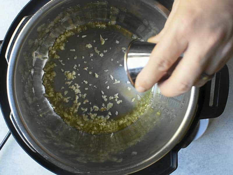 a hand holding a metal prep container pouring garlic and olive oil into an instant pot
