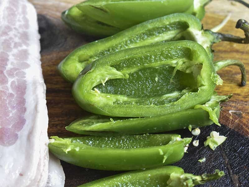 jalapeno peppers cut in half from step to tip with all seeds and white membrane removed.  Sitting on a brown wooden cutting board next to a stack of uncooked bacon strips.