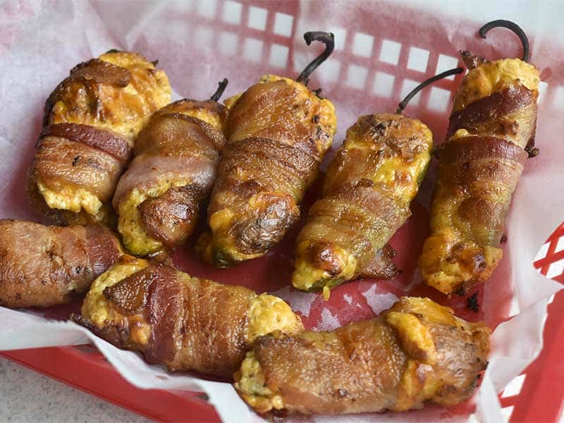 bacon wrapped smoked jalapeno poppers with cheddar and cream cheese stuffing sitting in a red food basket with white culinary paper lining it.