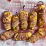 overhead photo of fully cooked bacon wrapped smoked jalapeno peppers with cheddar cheese and cream cheese filling in a red plastic food basket with white culinary paper