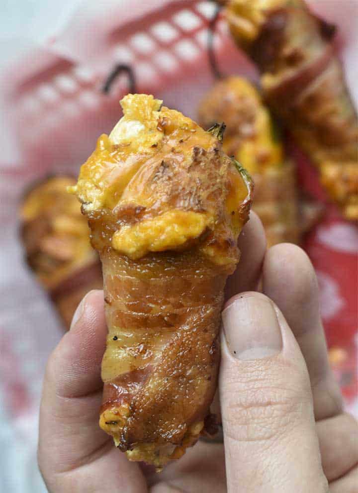 bacon wrapped smoked jalapeno popper with cheddar and creese filling.  Single cooked pepper being held above basket of other peppers in background.