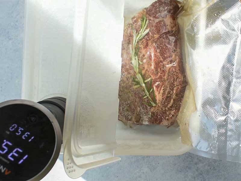 chuck roast in and rosemary in a vacuum sealer bag being lowered into a large plastic sous vide container filled with water.