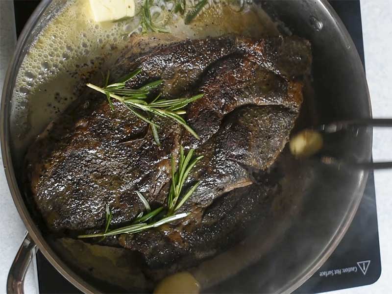 chuck roast being seared in a metal pan with fresh rosemary, butter and garlic cloves