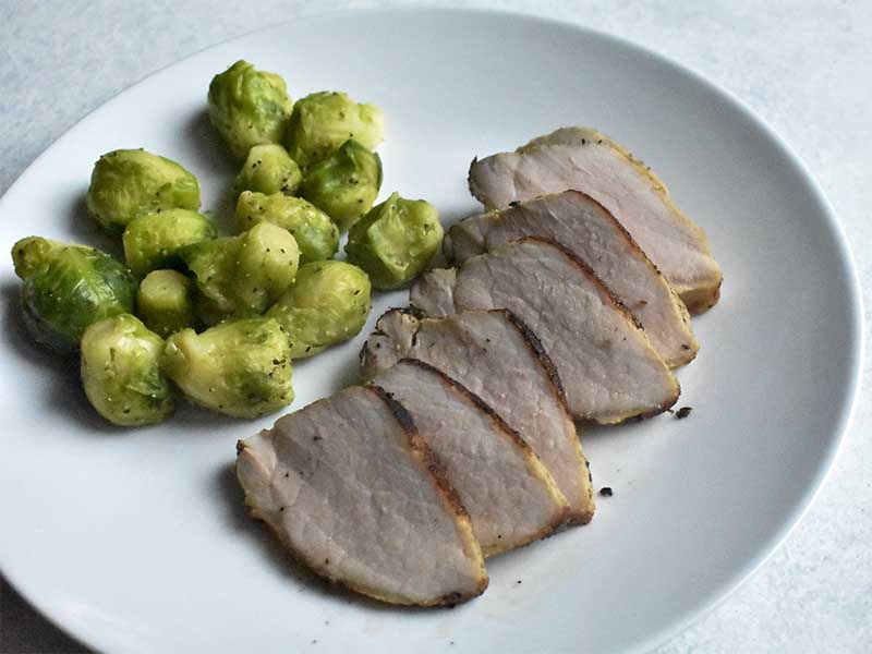sous vide pork tenderloin sliced sitting next to brussels sprouts on a white plate sitting on a white table