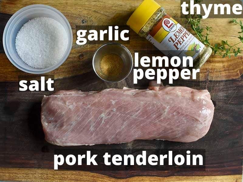 ingredients for sous vide pork tenderloin sitting on a wooden cutting board, labeled pork tenderloin, salt, garlic, lemon pepper and thyme