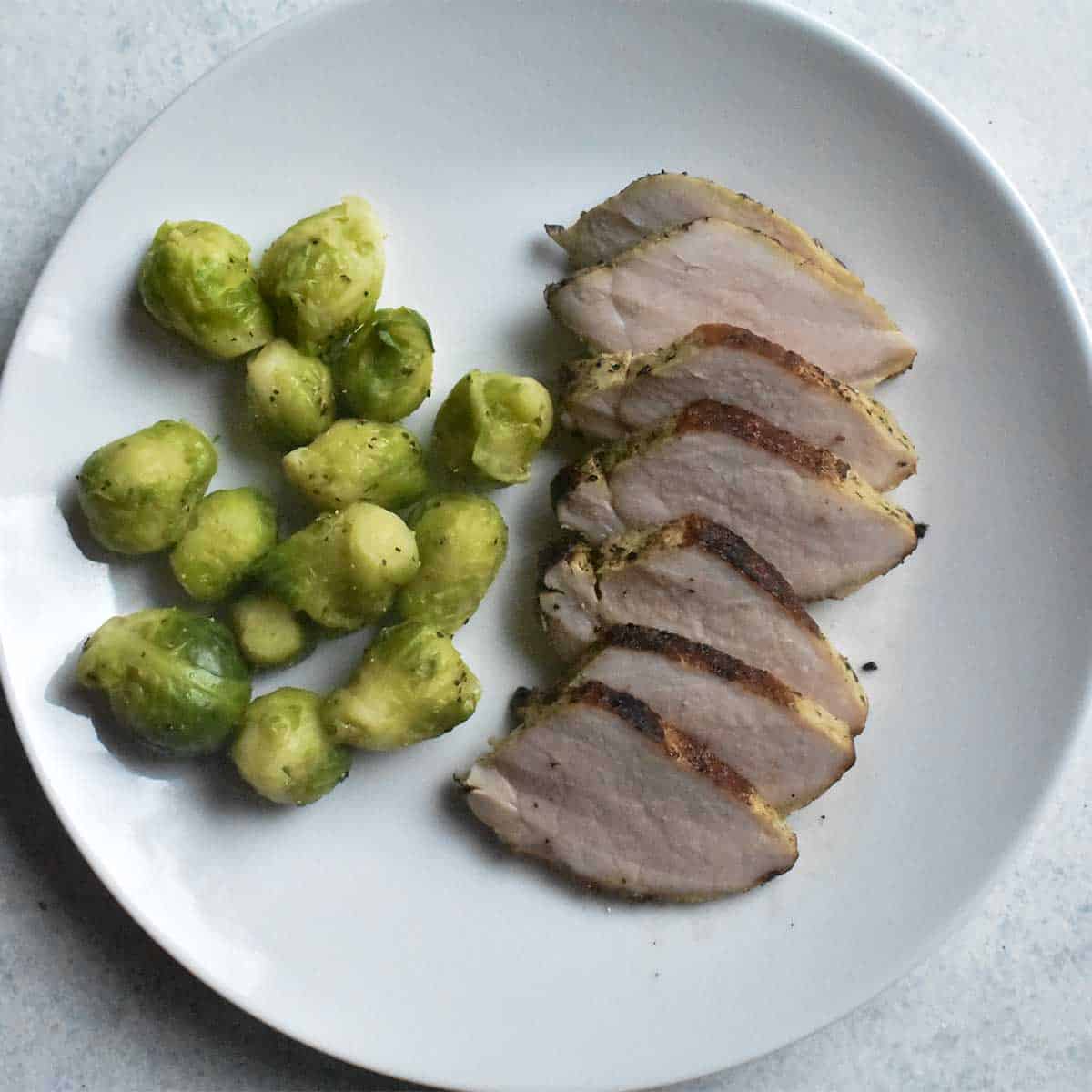 overhead shot of pork tenderloin sliced into medallions with brussels sprouts on a white plate sitting on a white table