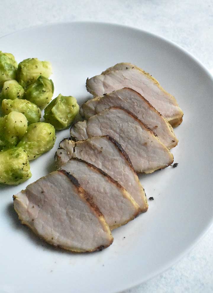Sous vide pork tenderloin sliced into medalions sitting on a white plate next to seasoned brussels sprouts