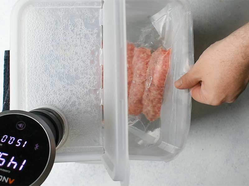 sausage in a plastic bag being lowerd into a sous vide water bath in a clear plastic container filled with water