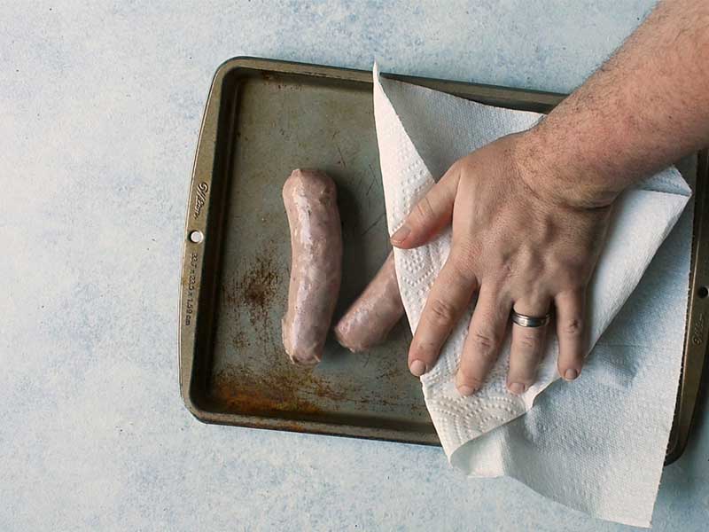 cooked sausage sitting on a brown metal baking pan. The sausage is being patted with paper towel by a hand reaching in from off camera. 