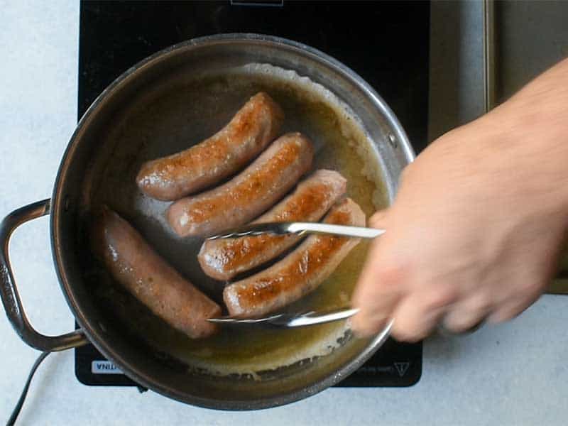 sausage being browned with butter in a silver pan on a black burner. A hand with tongs is reaching into the pan.