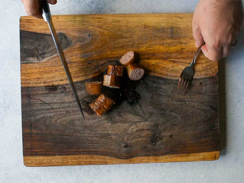 sausage cut into pieces on a cutting board. Hands are holding a knife and fork on either side of the sausage.