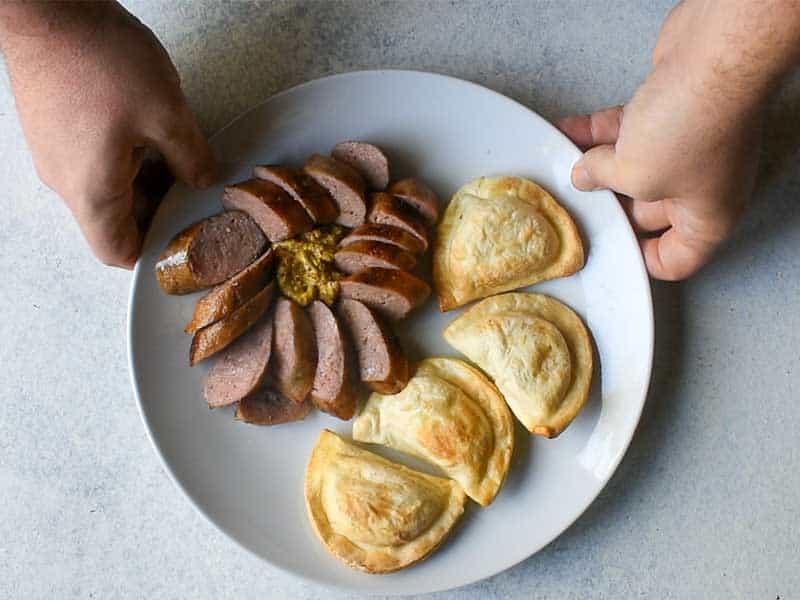 sliced sausage with dijon mustard and pirogi on a white plate being held by a hand on either side of the plate