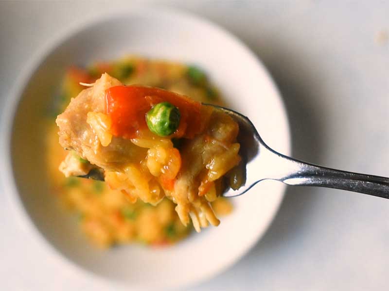 arroz con pollon on a metal fork above a white bowl of more chicken and yellow rice on a white table