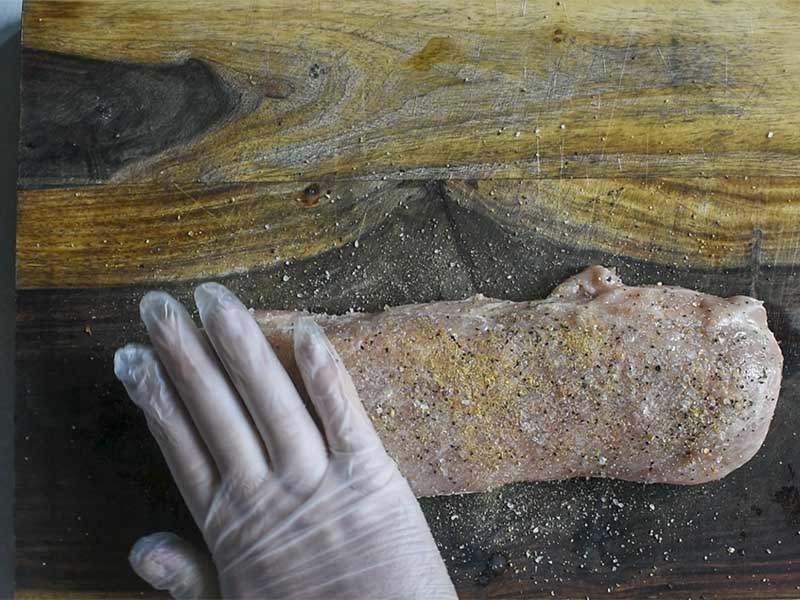 pork tenderloin seasoned with lemon pepper on a wooden board being patted with a gloved hand