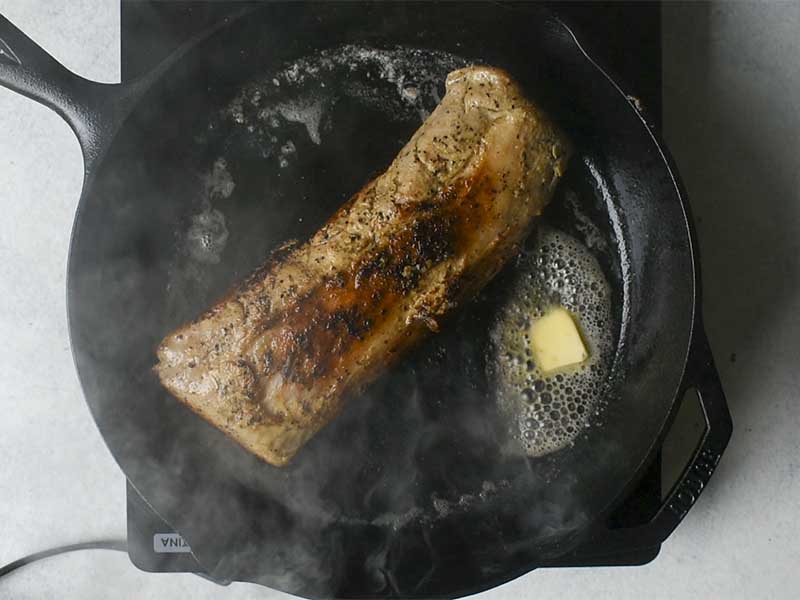 pork tenderloin being seared in a cast iron pan with butter melting alongside
