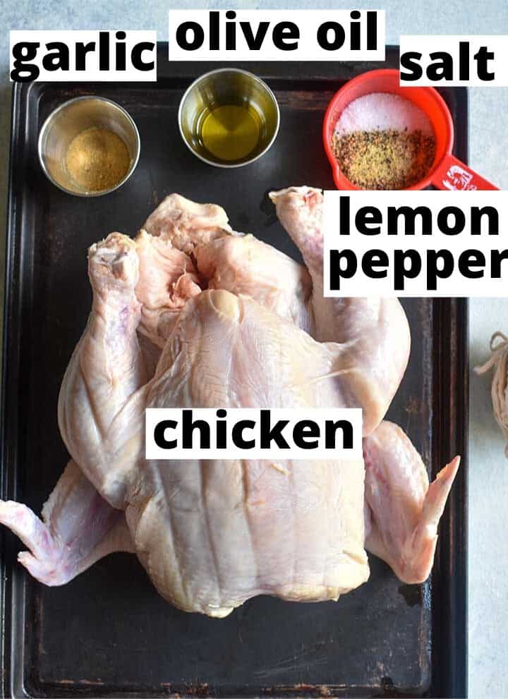recipe ingredients (garlic, olive oil, salt, lemon pepper and a whole chicken) sitting on a black baking tray on top of a white table.