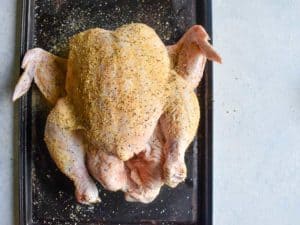 whole chicken seasoned with lemon pepper laying on a dark baking sheet.
