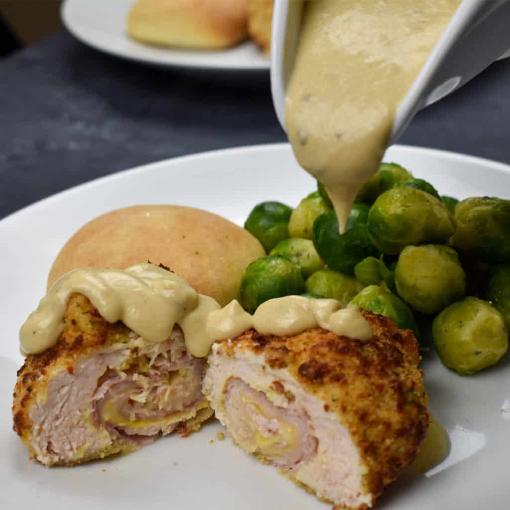 chicken cordon bleu with a dijon mustard sauce being poured overtop.  Brussels sprouts and a bread roll are also on the plate