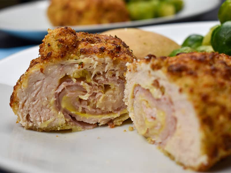 air chicken cordon bleu sliced open on a white plate with bread rolls and brussels sprouts