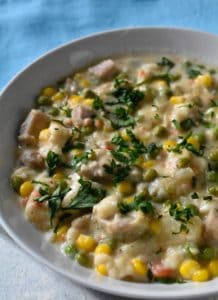 Chicken in dumplings topped with corn topped with parsley and sitting in a white bowl. There is a blue napkin in the background.