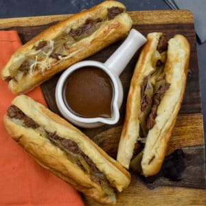 three french dip sandwiches on a wooden board with au jus sauce in a white dipping bowl