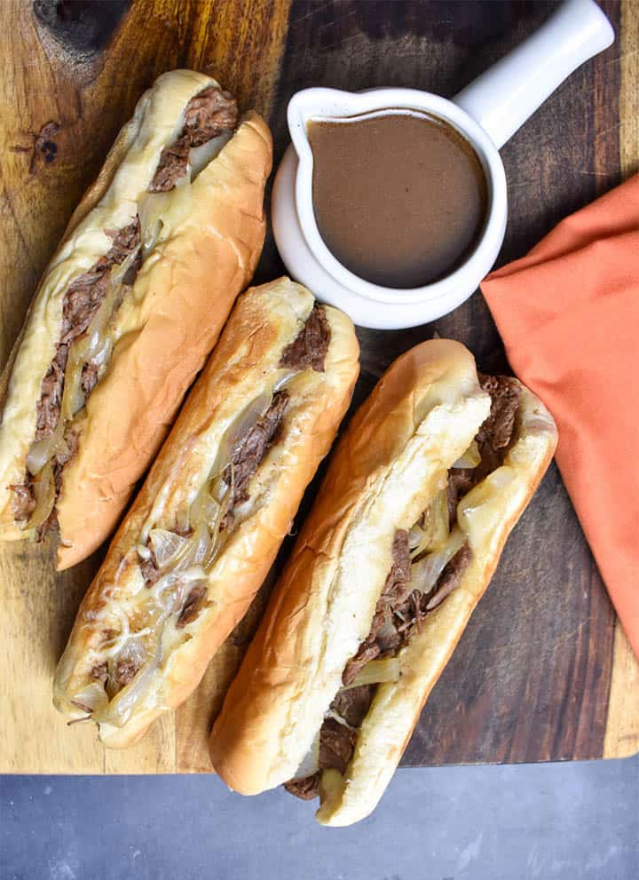 three french dip sandwiches with onions on a wooden cutting board with au jus dipping sauce in a white gravy bowl