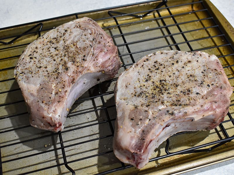 two pork chops which have been cooked in a sous vide but not yet seared.  Seasoned with salt and pepper and sitting on a wire rack above a baking sheet.