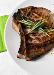 double bone pork chop sliced along the blone, sitting a white plate and topped with rosemary sprigs. There is a green napkin on a white table underneath.