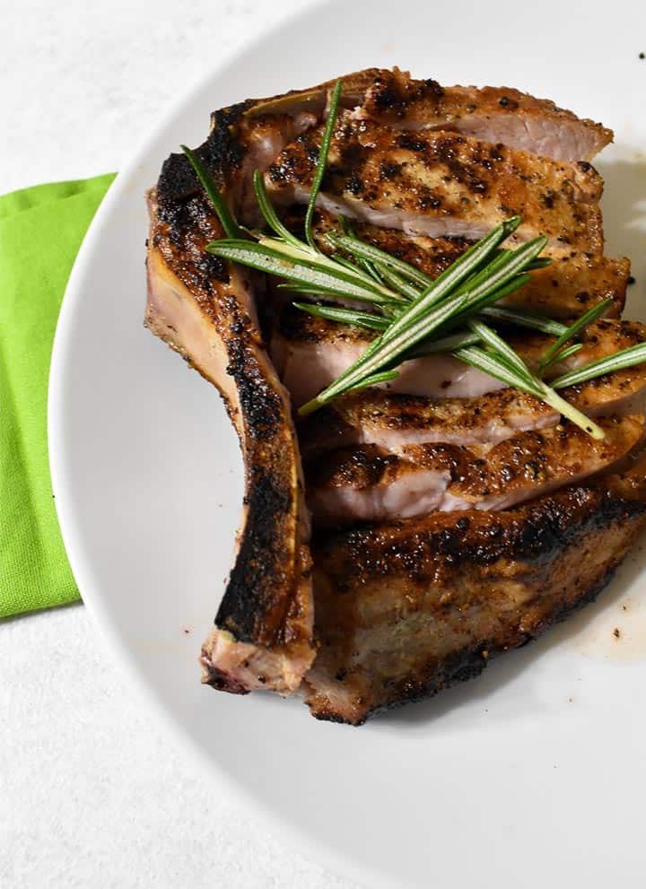 double bone pork chop sliced along the blone, sitting a white plate and topped with rosemary sprigs.  There is a green napkin on a white table underneath.