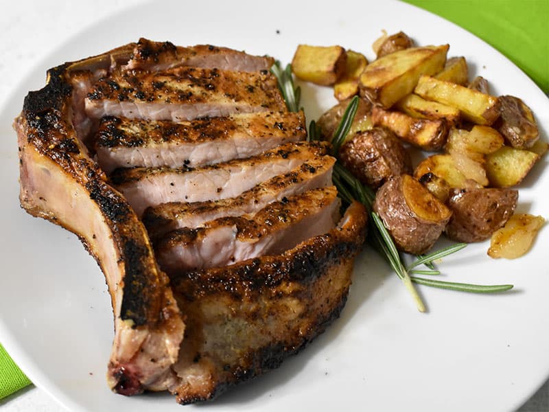 pork chop sliced alongside the bone on a white plate with roasted potatoes alongside.