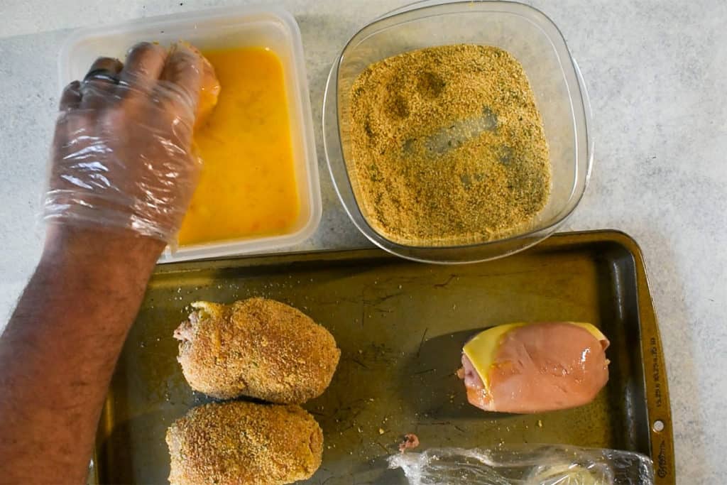 rolled and stuffed chicken breast being breaded by dunking in egg next to a tupperware container container breadcrumbs