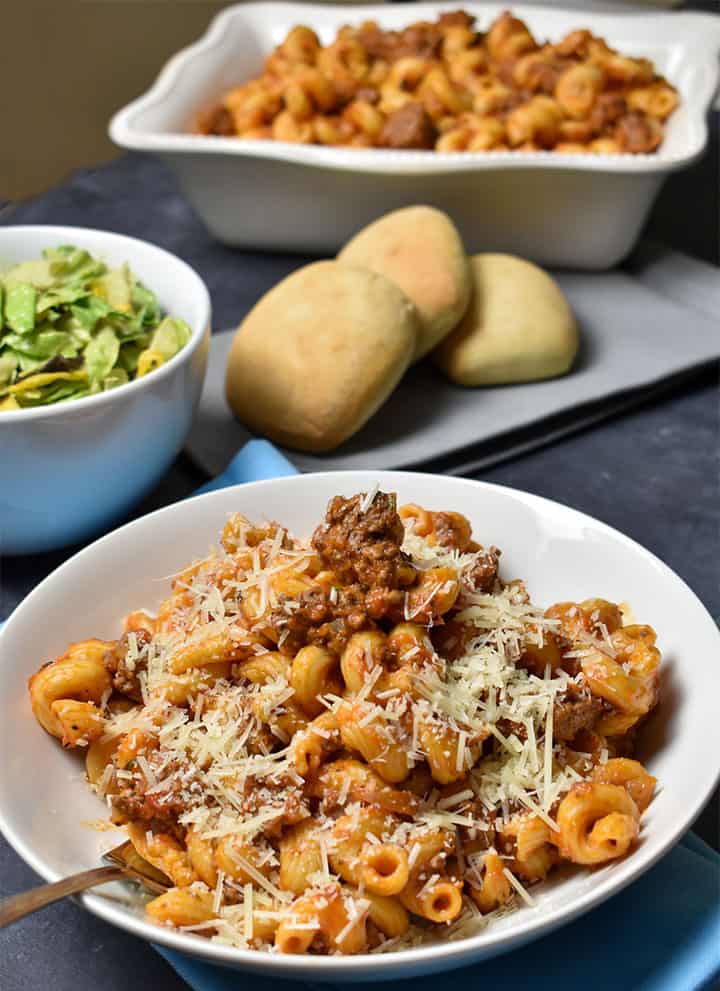 Goulash in a white bowl on a black table with rolls and salad