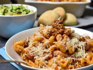 noodles with ground beef and tomato sauce in a whit ebowl with salad and rolls in the background