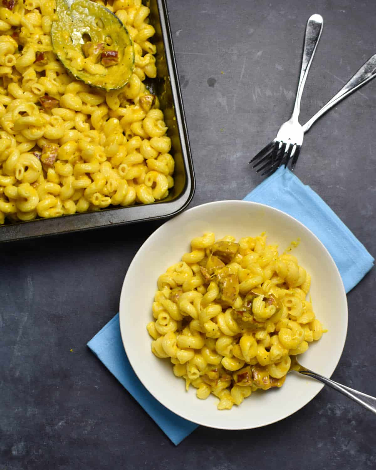 overhead shot of mac and cheese with hot dogs. There is a serving of mac and cheese in a white bowl in the lower right corner, with the remaining mac and cheese in a pan in the upper left corner. There is one fork in the bowl and two more resting on the table.