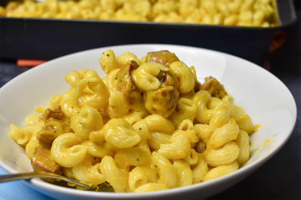 a white bowl filled with mac and cheese with a fork. there is a pan with more mac and cheese in the background